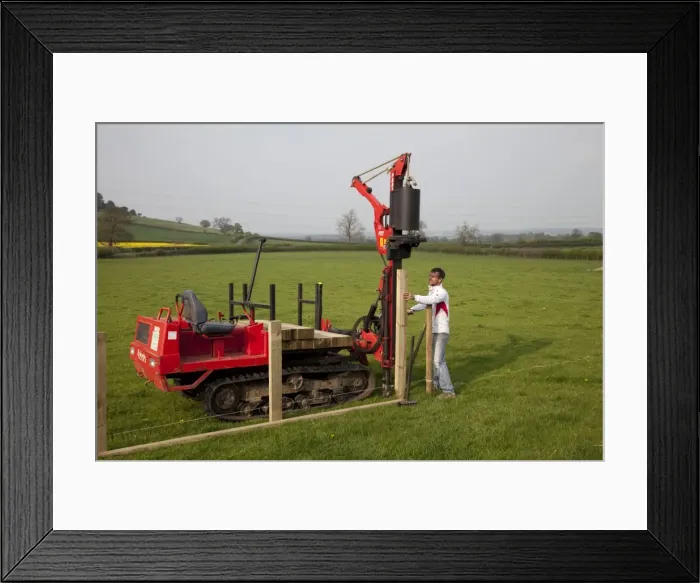 Framed Print : Workman driving timber posts into ground for new post and rail field fence with dedicated Kubota Protech tracked Post Rammer - Cotswolds - UK