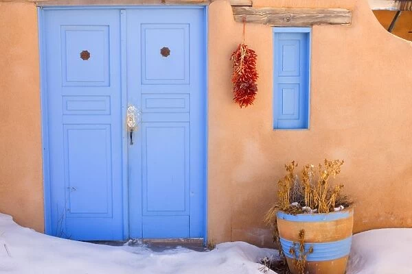 Adobe style house - simple beige coloured house in adobe style with a bright blue coloured door and a string of dried chilly peppers, called ristra, hanging from a beam. In winter - Rancho de Taos, New Mexico, USA