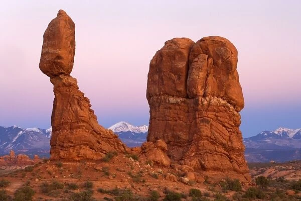 Balanced rock - sandstone rock formation in the shape of a spire with an oval shaped rock balancing on top of it - snow covered Manti-La Sal Mountains and the Windows Section of the park with its numerous arches is visible in the background - at