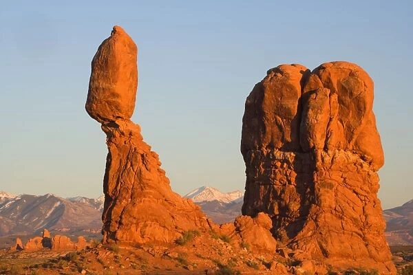 Balanced rock - sandstone rock formation in the shape of a spire with an oval shaped rock balanching on top of it. The snow covered Manti-La Sal Mountains and the Windows Section of the park with its numerous arches is visible in the background