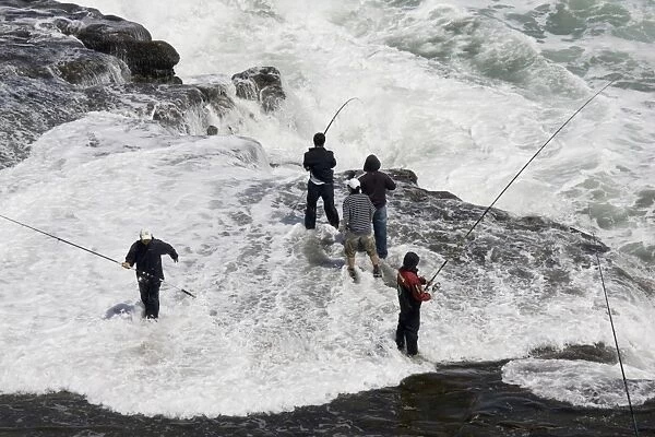 Men fishing on rock outcrop amidst breaking waves