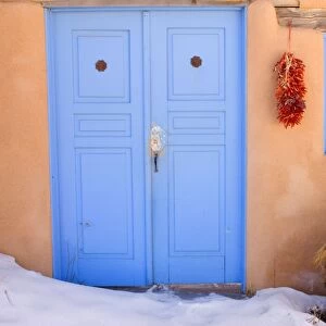 Adobe style house - simple beige coloured house in adobe style with a bright blue coloured door and a string of dried chilly peppers, called ristra, hanging from a beam. In winter - Rancho de Taos, New Mexico, USA